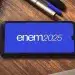 Celular na mesa exibindo o logo ENEM 2025 em fundo azul, com caderno e caneta ao lado