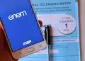 Mão segurando celular com tela do aplicativo ENEM aberto e caderno azul de prova do ENEM ao fundo