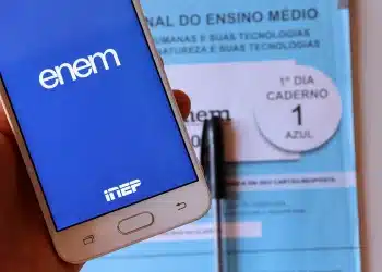 Mão segurando celular com tela do aplicativo ENEM aberto e caderno azul de prova do ENEM ao fundo
