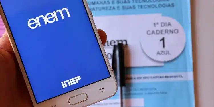 Mão segurando celular com tela do aplicativo ENEM aberto e caderno azul de prova do ENEM ao fundo