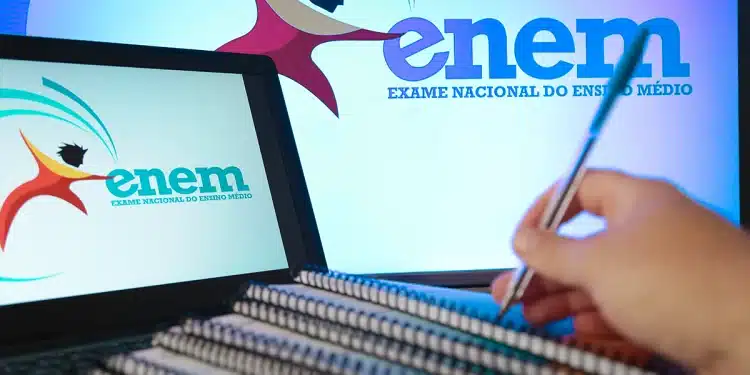 Estudante revisando cadernos com caneta ao lado de notebook exibindo a logo do Enem (Exame Nacional do Ensino Médio), preparando-se para a prova.