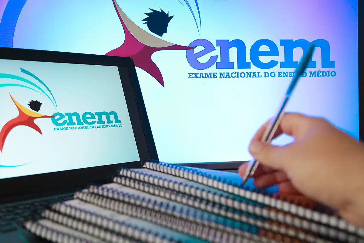 Mão escrevendo em caderno com logos do ENEM em tela de computador e monitor ao fundo