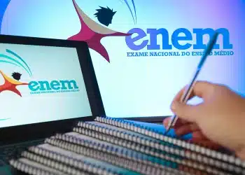 Pessoa escrevendo com caneta diante de notebooks com logo do ENEM e cadernos organizados para estudo.