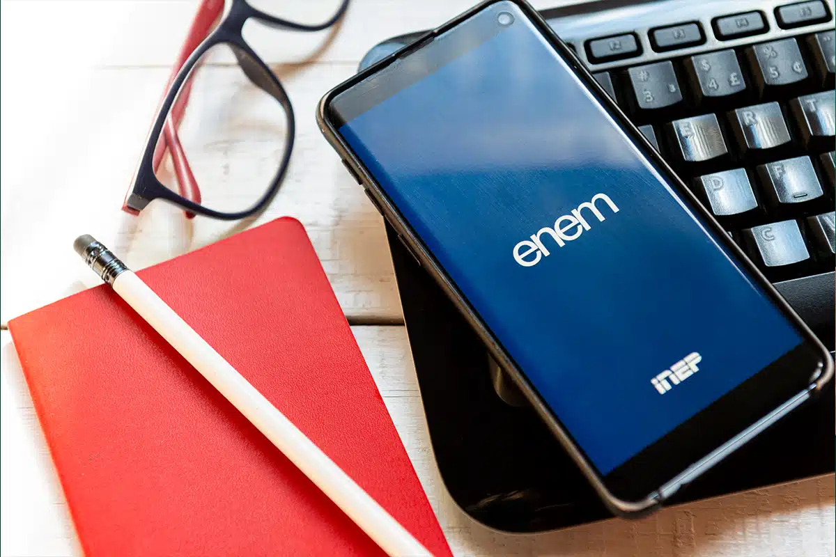 Tela de celular com a logo do Enem e Inep, simbolizando a consulta do Cartão de Confirmação na Página do Participante, ao lado de óculos e caderno de anotações.