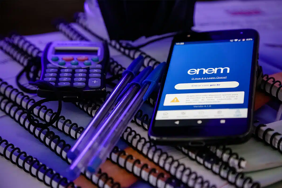 Smartphone com aplicativo ENEM aberto, ao lado de canetas azuis, calculadora e cadernos em ambiente de estudo