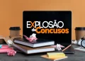 Mesa com livros, laptop mostrando Explosão de Concursos e materiais de estudo espalhados.