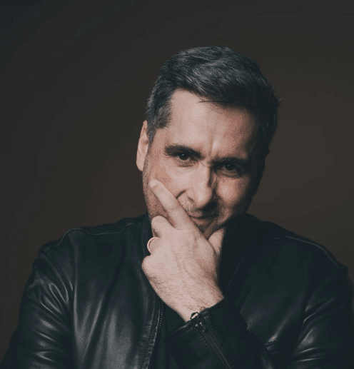 Homem com cabelo grisalho e jaqueta preta fazendo gesto de mão no rosto, fundo escuro