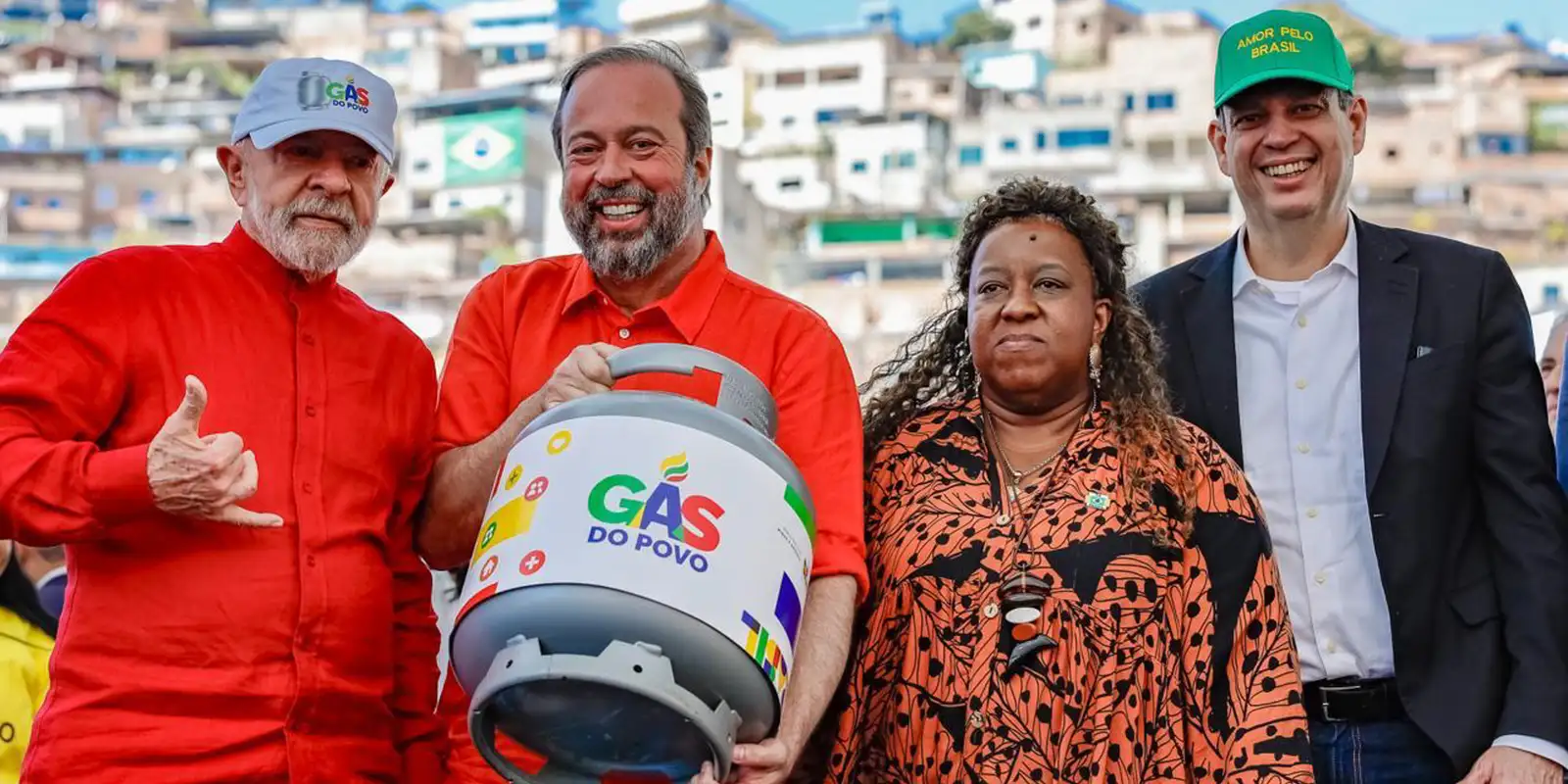 Quatro pessoas, duas com camisas vermelhas, segurando botijão de gás do projeto Gás do Povo em comunidade
