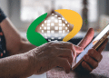 Logo do INSS sobre as mãos de uma pessoa idosa utilizando um celular para consultar o calendário de pagamento de benefícios e auxílios, referente a outubro e novembro.