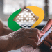 Logo do INSS sobre as mãos de uma pessoa idosa utilizando um celular para consultar o calendário de pagamento de benefícios e auxílios, referente a outubro e novembro.