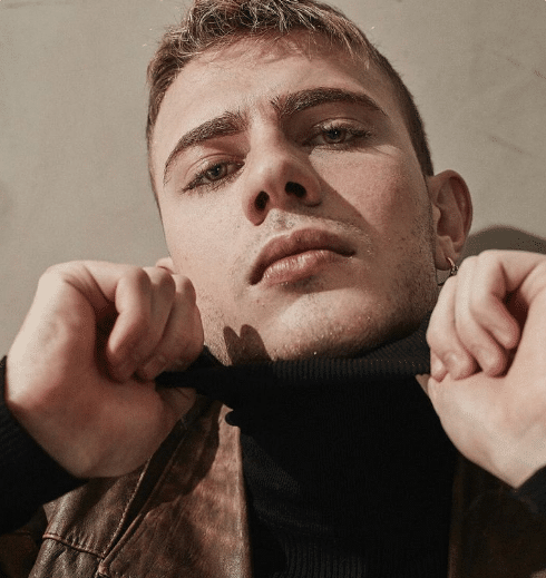 Homem jovem segurando a gola do suéter preto com expressão séria e jaqueta de couro marrom