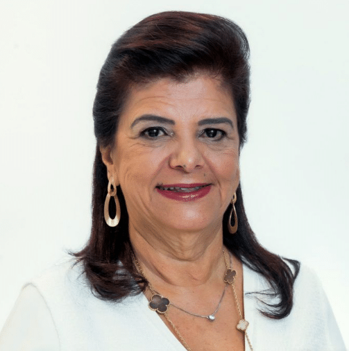 Mulher madura sorrindo, com cabelo preso, usando blusa branca e acessórios dourados