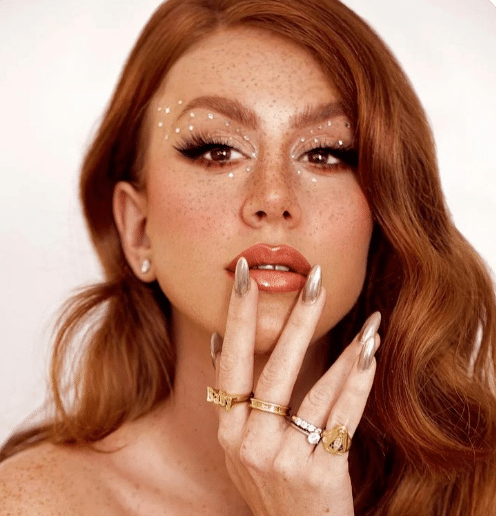Mulher ruiva com maquiagem artística, glitter branco ao redor dos olhos e unhas metálicas douradas