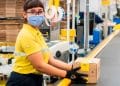 Funcionária do Mercado Livre em centro de distribuição, utilizando máscara e luvas, segurando uma caixa em esteira de logística.