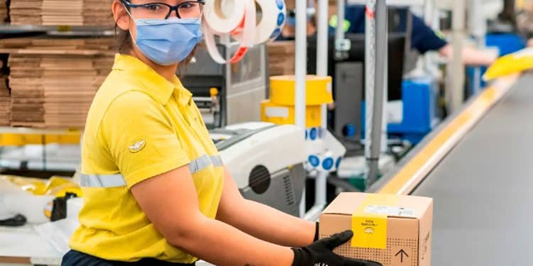 Funcionária do Mercado Livre em centro de distribuição, utilizando máscara e luvas, segurando uma caixa em esteira de logística.