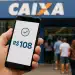 Pessoa segura celular com valor de R$108 em frente à agência da Caixa