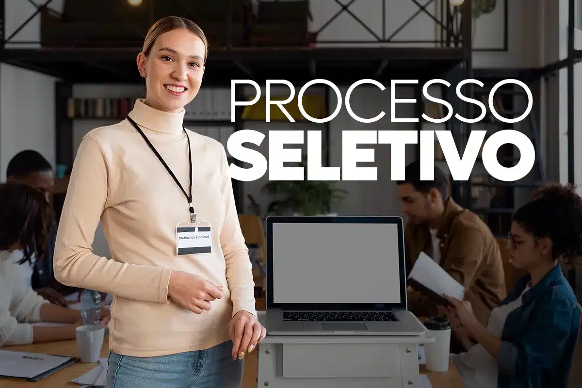 Jovem sorridente com crachá em frente a um notebook, representando a estudante que atende aos requisitos do Processo Seletivo do INSS, como matrícula em ensino médio ou superior.
