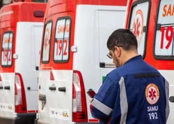 Homem com uniforme do SAMU 192 verifica celular próximo a fila de ambulâncias brancas e vermelhas estacionadas.