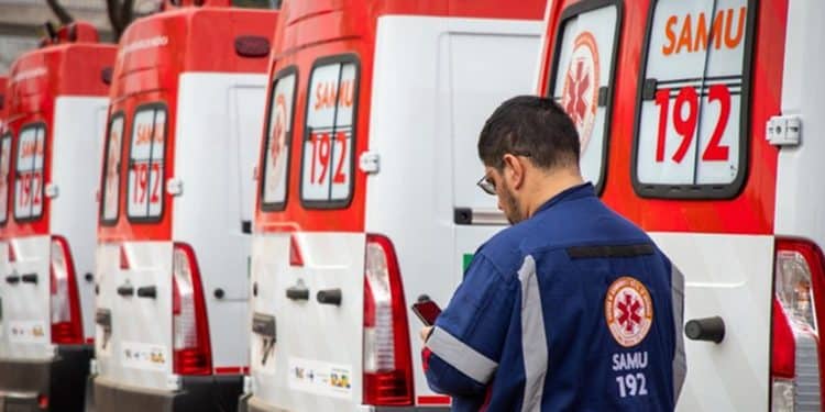 Homem com uniforme do SAMU 192 verifica celular próximo a fila de ambulâncias brancas e vermelhas estacionadas.