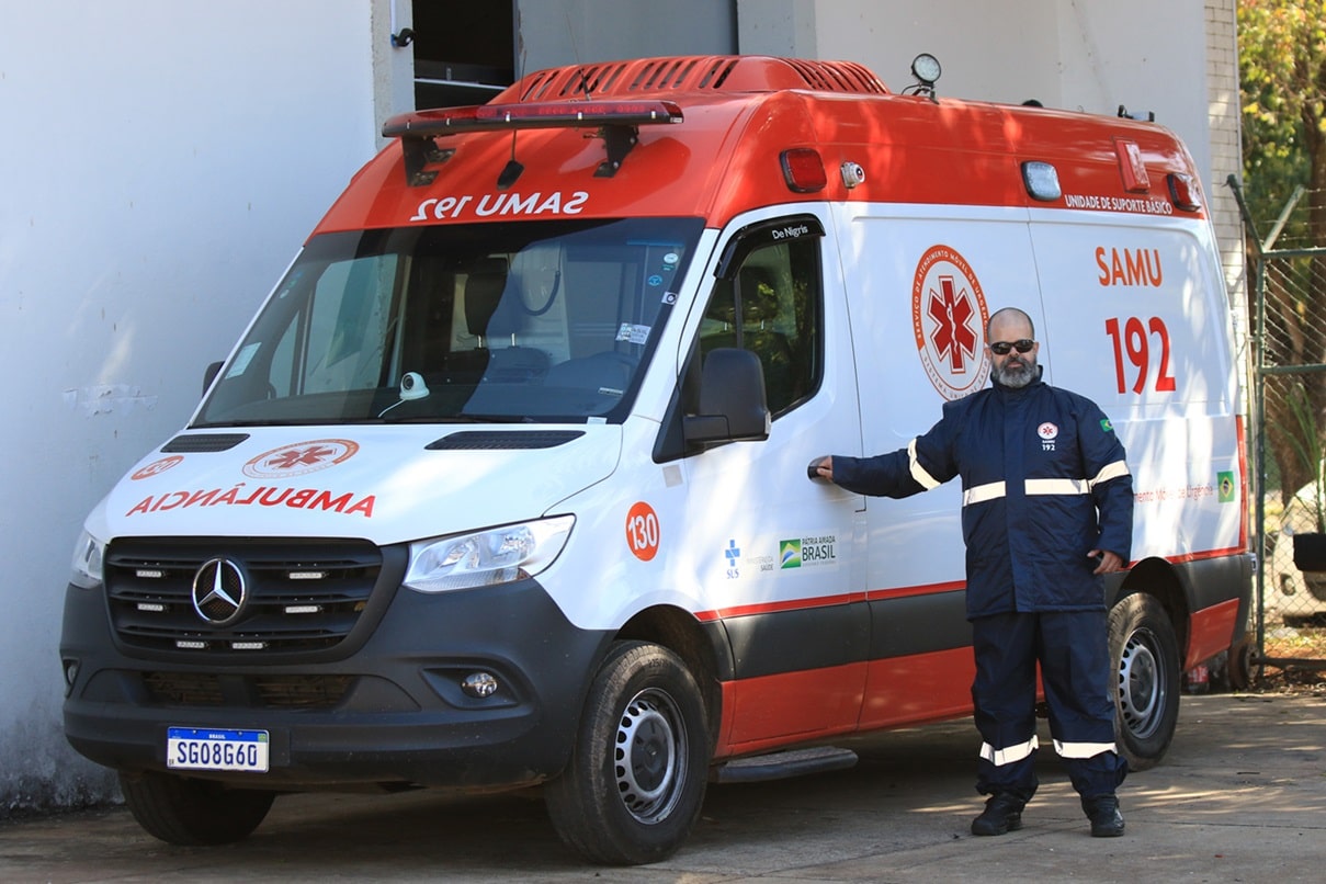 Ambulância SAMU 192 branca e vermelha com profissional de saúde de uniforme azul escuro ao lado.