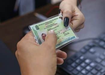 Mãos segurando a Carteira de Identidade Nacional brasileira com foto e dados pessoais durante processo de emissão no mutirão gratuito