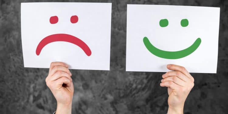Mãos segurando cartões com rostos de emoções: um rosto triste vermelho e um rosto feliz verde. Ilustra a dualidade de sentimentos entre ser engraçado e estar triste.