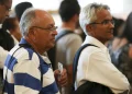 Grupo de pessoas em fila, incluindo homens com cabelo grisalho e óculos, esperando em ambiente fechado