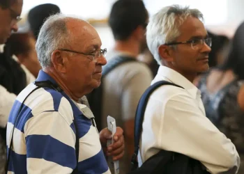 Grupo de pessoas em fila, incluindo homens com cabelo grisalho e óculos, esperando em ambiente fechado