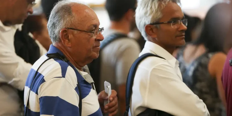 Grupo de pessoas em fila, incluindo homens com cabelo grisalho e óculos, esperando em ambiente fechado