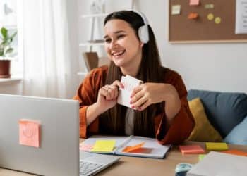 Mulher trabalhando em home office, sorrindo enquanto segura um cartão de notas e utilizando fones de ouvido, com computador e post-its ao fundo.