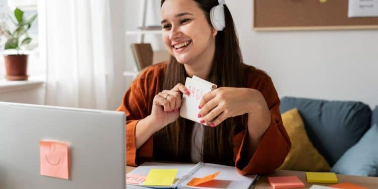 Mulher trabalhando em home office, sorrindo enquanto segura um cartão de notas e utilizando fones de ouvido, com computador e post-its ao fundo.