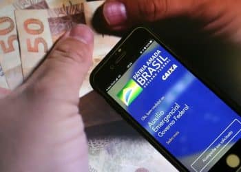 Mão segurando smartphone com o aplicativo do Auxílio Emergencial da Caixa aberto, com notas de R$ 50 reais ao fundo, ilustrando a notificação de devolução do benefício.