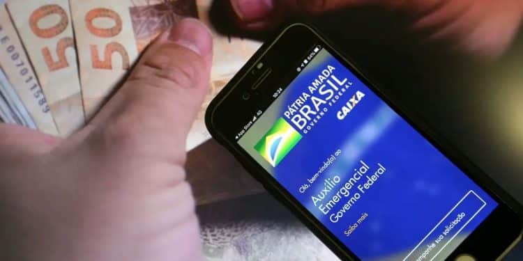 Mão segurando smartphone com o aplicativo do Auxílio Emergencial da Caixa aberto, com notas de R$ 50 reais ao fundo, ilustrando a notificação de devolução do benefício.