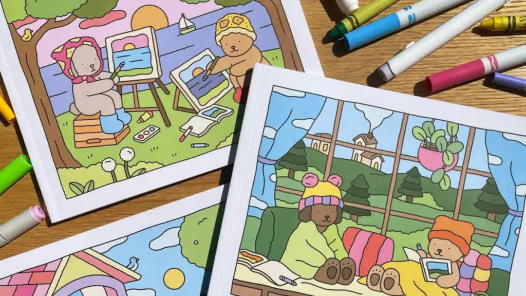 Livros de colorir abertos com ilustrações adoráveis de ursinhos e cachorrinhos em cenas tranquilas, rodeados por canetas e lápis de cor, destacando a arte da Bobbie Goods.