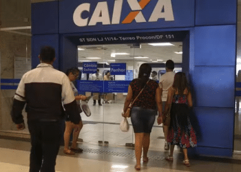 Pessoas entrando na agência Caixa Econômica Federal com portas de vidro e fachada azul com logo da Caixa.