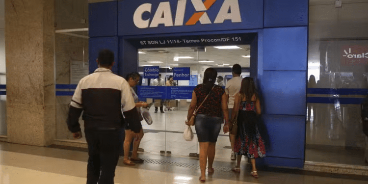 Pessoas entrando na agência Caixa Econômica Federal com portas de vidro e fachada azul com logo da Caixa.
