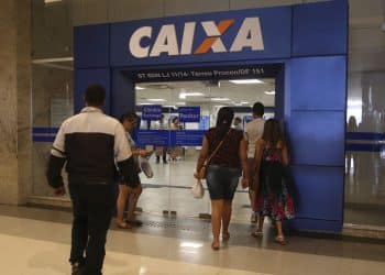 Entrada da agência da Caixa Econômica com pessoas entrando pela porta de vidro em área interna.