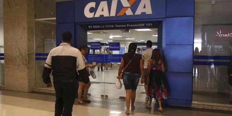 Entrada da agência da Caixa Econômica com pessoas entrando pela porta de vidro em área interna.