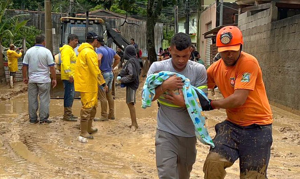 Homem carregando bebê enrolado em cobertor com socorrista em rua inundada e cheia de lama