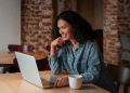 Mulher com cabelo cacheado sorrindo enquanto utiliza notebook em um café com parede de tijolos
