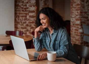 Mulher com cabelo cacheado sorrindo enquanto utiliza notebook em um café com parede de tijolos
