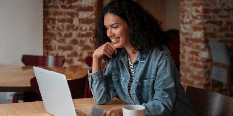 Mulher com cabelo cacheado sorrindo enquanto utiliza notebook em um café com parede de tijolos