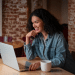 Mulher com cabelo cacheado sorrindo enquanto utiliza notebook em um café com parede de tijolos