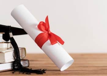 Diploma universitário enrolado com laço vermelho, capelo de formatura e livros empilhados. A imagem simboliza a conclusão de cursos que levam ao sucesso em concursos e na carreira pública.