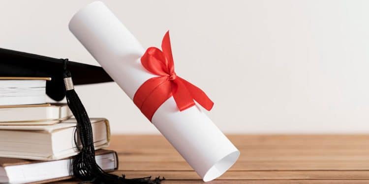 Diploma universitário enrolado com laço vermelho, capelo de formatura e livros empilhados. A imagem simboliza a conclusão de cursos que levam ao sucesso em concursos e na carreira pública.