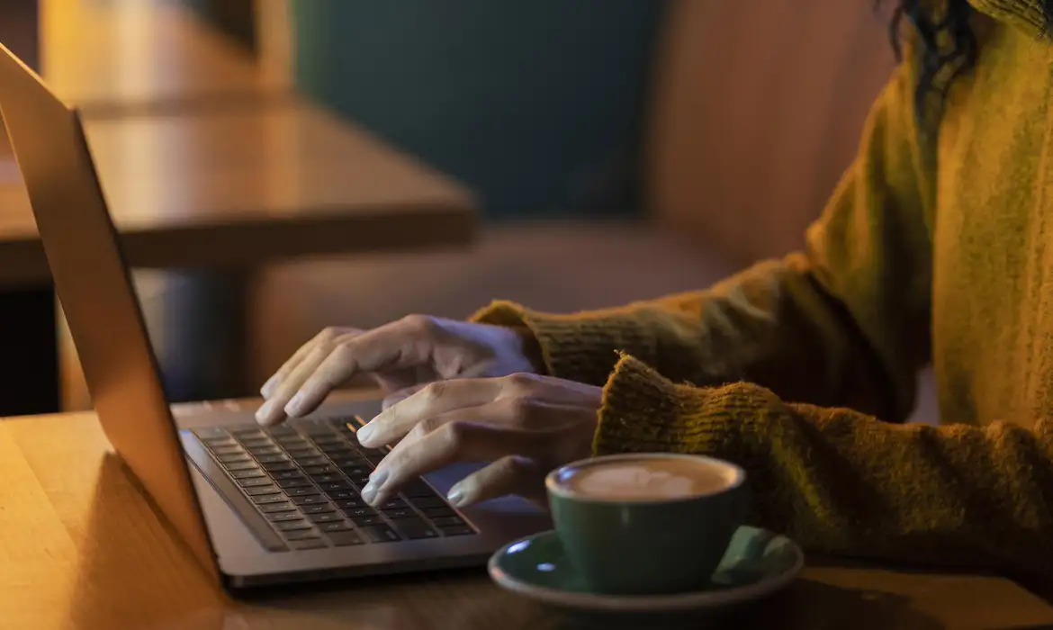 Mãos de pessoa usando laptop em ambiente aconchegante com xícara de café ao lado