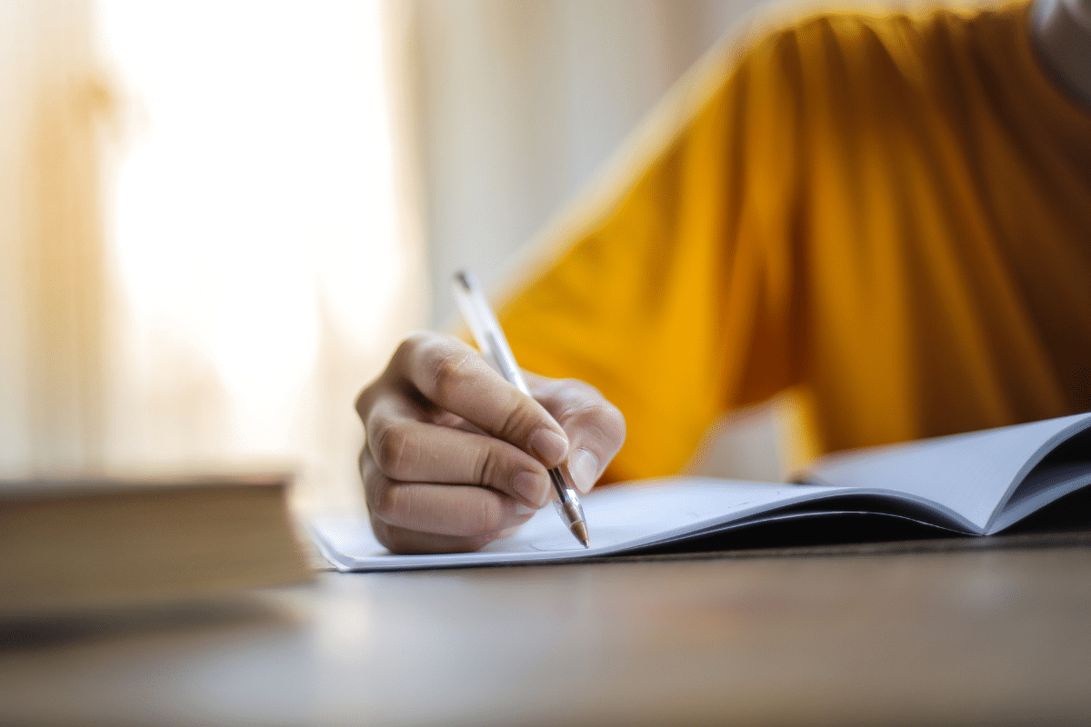 Mão segurando caneta transparente escrevendo em um caderno aberto sobre a mesa ao lado de um livro fechado