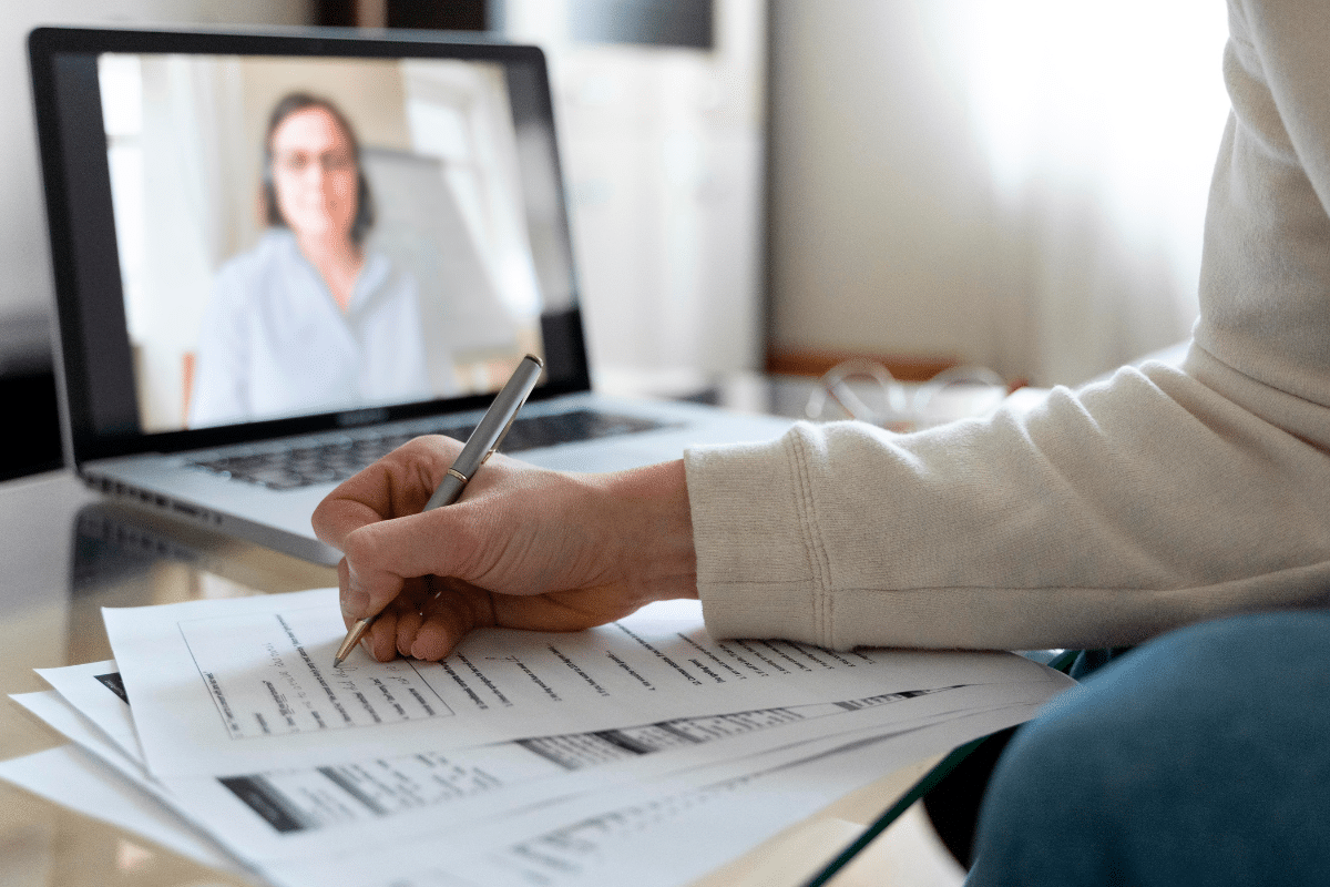 Pessoa escrevendo em documentos durante chamada de vídeo em laptop com consultora ao fundo