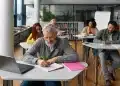Grupo de adultos sentados em mesas em uma sala de aula moderna, focados em estudar e escrever em cadernos