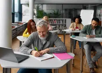 Grupo de adultos sentados em mesas em uma sala de aula moderna, focados em estudar e escrever em cadernos